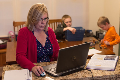 woman on laptop on kitchen counter with two boys playing in background