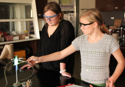 An Advancement Placement science student shows a young student how to work in the lab. Both wear safety goggles.