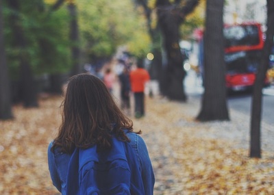 A child shown from behind with a backpack walks down a city street with fall leaves all around