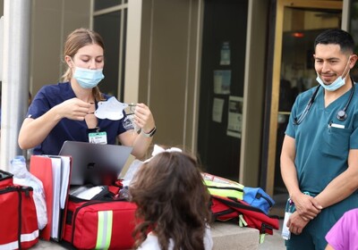 A nurse and a doctor hand a face mask to a child, outside.