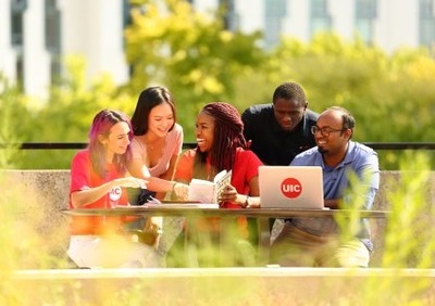 A diverse group of enthusiastic students collaborate outside around a laptop with a University of Illinois Chicago sticker on the lid