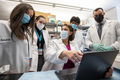 several masked, white coat lab workers gathered around a laptop in lab