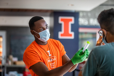 Masked Black male with orange SHIELD t-shirt h gloves handing test tube to man